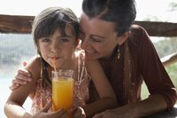 Girl uses a straw to drink a glass of orange juice next to her mother.