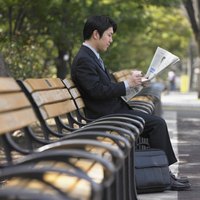 A businessman is reading a newsletter on a park bench.