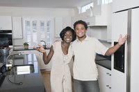 A smiling couple is standing in the kitchen by the refrigerator.