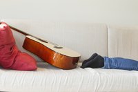 A man wearing dark socks and jeans lounges on the couch next a guitar and pillow.