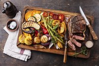 Grilled red meat and vegetables on a cutting board with seasonings.
