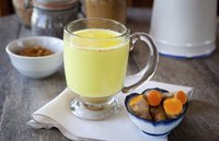 A hot drink made with turmeric, milk, honey, cinnamon and ginger next to a small bowl of fresh turmeric root.