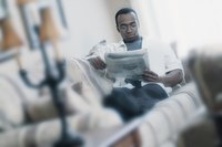 A man reclines on the couch while perusing the newspaper.
