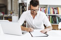 A young man sits down at his desk with his laptop and his note pad to begin writing his project report.