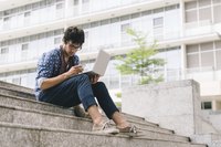 A writer sitting on a campus stairwell outside with a laptop and smartphone.