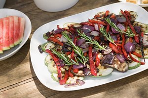 Plato de vegetales grillados con romero y recipiente con sandía, en una mesa en el patio.