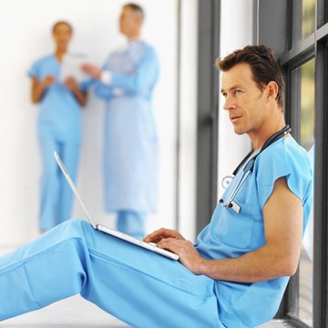 Close-up of a mid adult surgeon sitting on the floor working on laptop with two other medical personnel in background