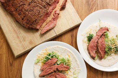 Grilled flank steak on wood cutting board next to two plates of tacos.