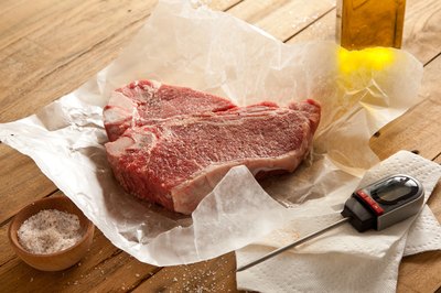 Raw steak on butcher paper next to thermometer, olive oil and salt.