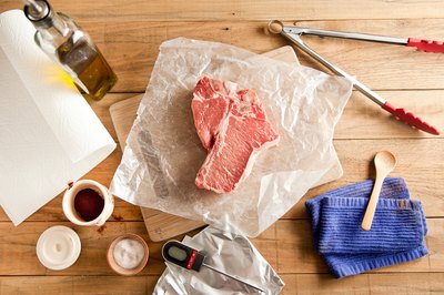 Raw steak on butcher paper with dry spices, tongs, thermometer, olive oil and utensils.