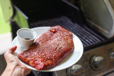 Seasoned skirt steak on a plate next to a BBQ grill.