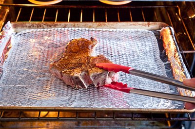 Steak on broiler pan in oven turned over by tongs.