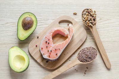 An overhead view of healthy dietary fats including salmon, avocado, nuts and seeds on a cutting board.