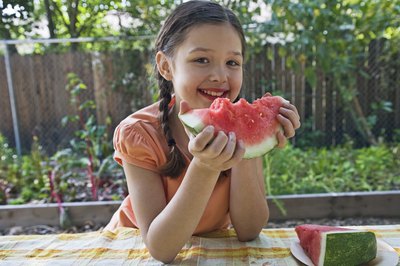 A little girl eating a slice of watermelon at an outdoor table near the garden.