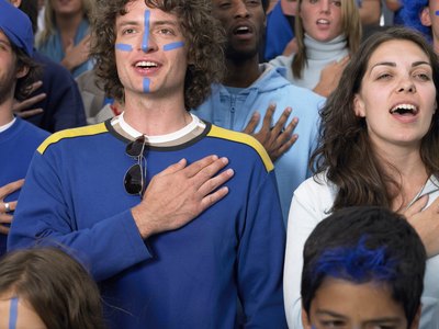 A young couple stand together while singing the national anthem, each has a hand placed over the heart