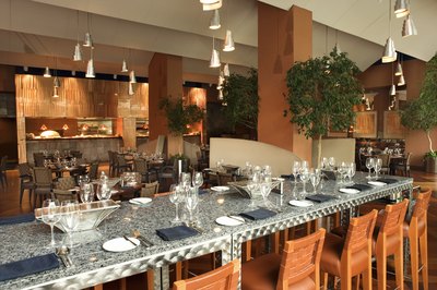 An interior of a restaurant shows a high marble bar with wooden chairs to offer a casual but sleek aesthetic