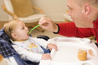 A man spoon feeds a baby in a highchair.