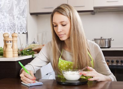 A woman weighs food on a scale while taking notes in her kitchen.
