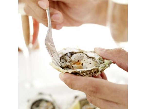 A close-up of a woman using a fork to loosen a raw oyster from the shell