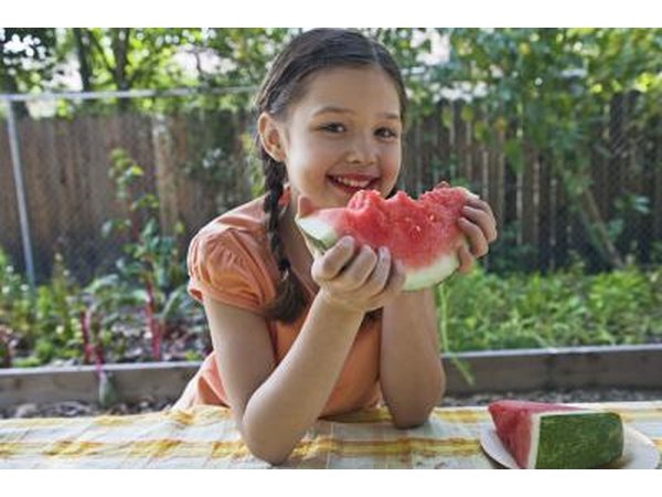 A little girl eating a slice of watermelon at an outdoor table near the garden.