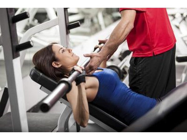 A woman does bench presses with the help of a trainer.