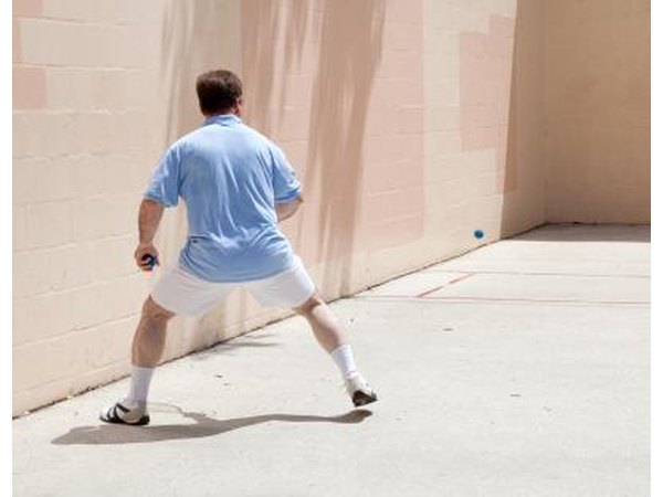 A man takes a wide squatted stance while playing racquetball outside.