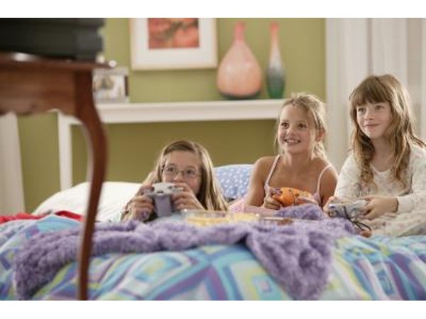Three girls playing a video game in bed while snacking on popcorn.