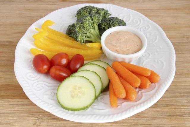 Slices of cucumber, carrot, yellow peppers, and broccoli on a plate with cherry tomatoes and a side of dressing.