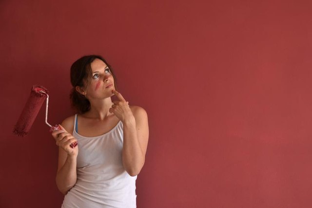 A woman holds a paint roller thinking in front of a red wall.