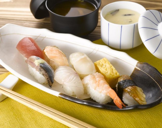 A platter of sushi on a placemat with chopsticks and soups.