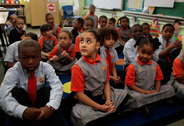Elementary students wearing school uniforms sit on a rug in the classroom. Elementary students wearing school uniforms sit on a rug in the classroom.
