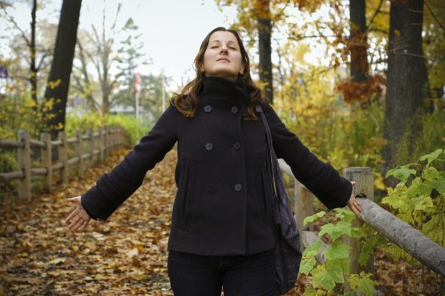 A woman inhaling a deep breath of fresh air on a walking trail in the woods on a fall day.