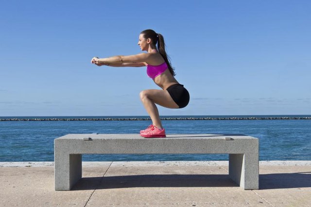 A woman doing prisoner squats on a bench near the water.