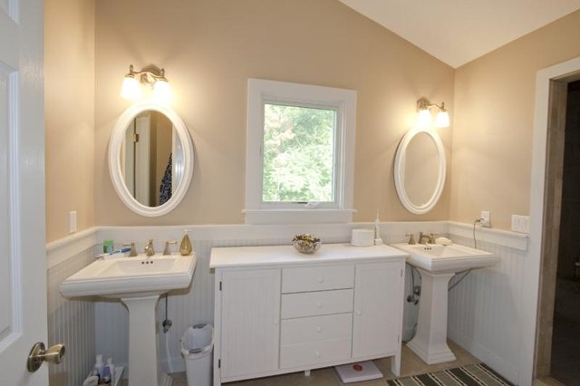 A white cabinet and window between two porcelain pedestal sinks in a bathroom.
