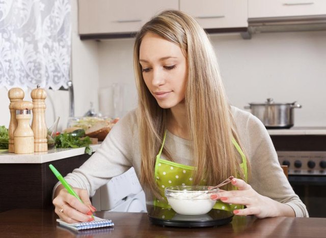 A woman weighs food on a scale while taking notes in her kitchen.