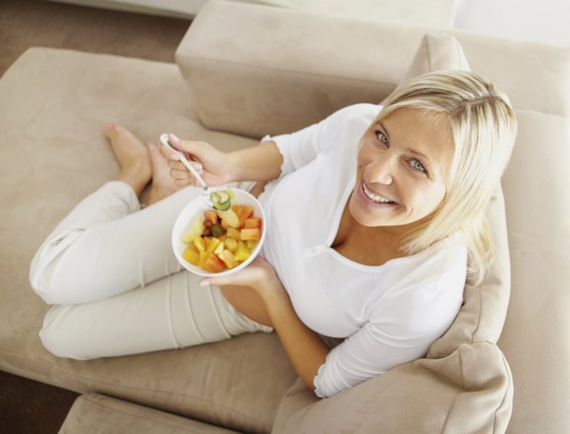 A woman reclines while eating a bowl of fruit on an ultra suede couch.