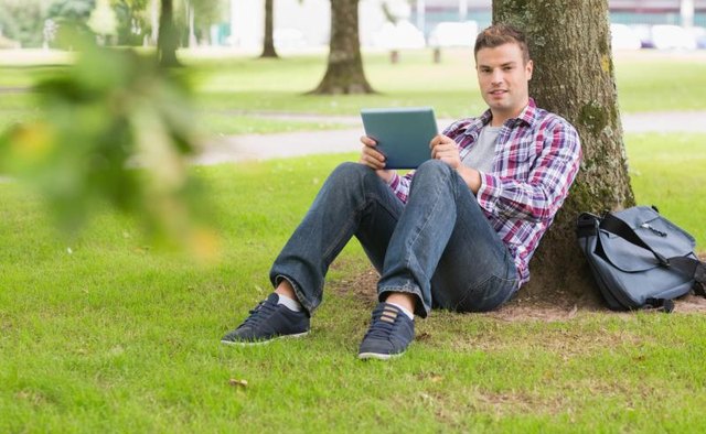 A leather messenger bag beside man leaning against tree.