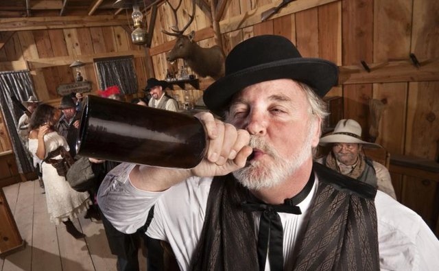 A man in Wild West dress drinking from a bottle, in front of others wearing Wild West costumes.