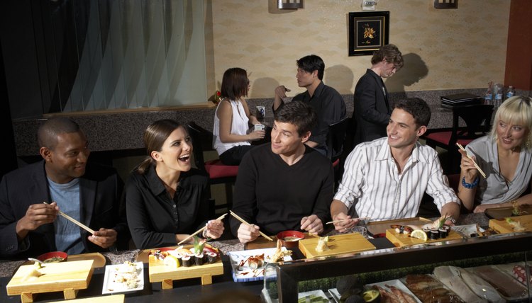 Five people sitting at counter in sushi bar, others in background, woman laughing