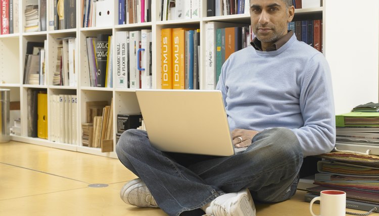 Man leaning against bookshelf, laptop computer on lap, portrait