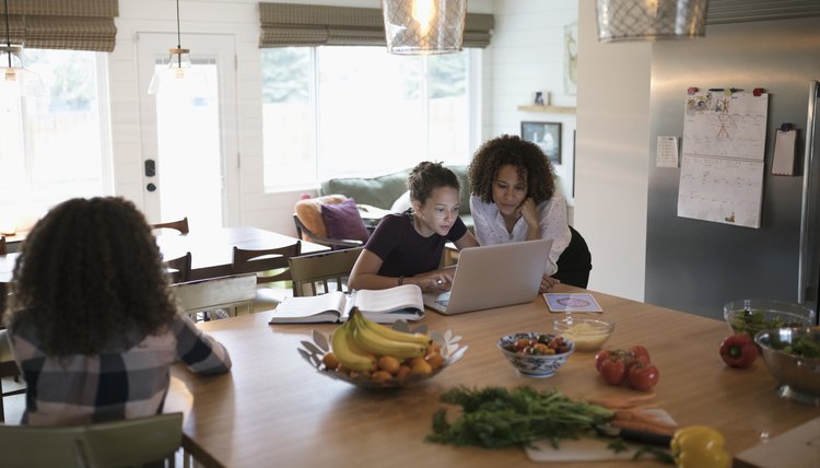 Mother helping tween daughter doing homework at laptop in kitchen