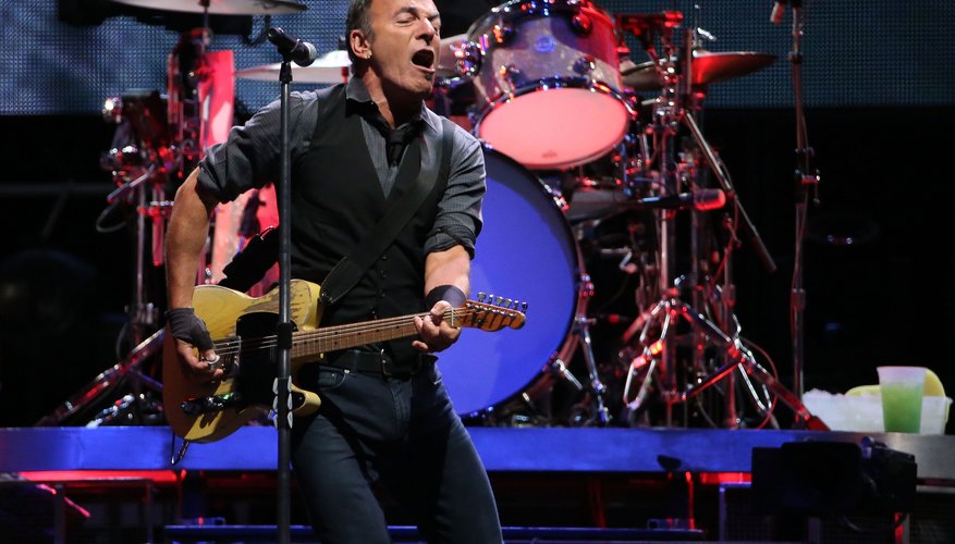 Bruce Springsteen singing into the mic while playing the guitar on stage. Bruce Springsteen singing into the mic while playing the guitar on stage.