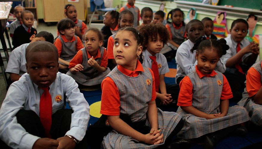 Elementary students wearing school uniforms sit on a rug in the classroom. Elementary students wearing school uniforms sit on a rug in the classroom.
