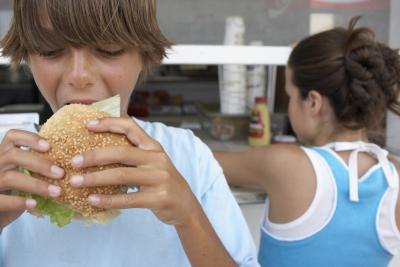 A boy eats a hamburger in front of a take-out stand.