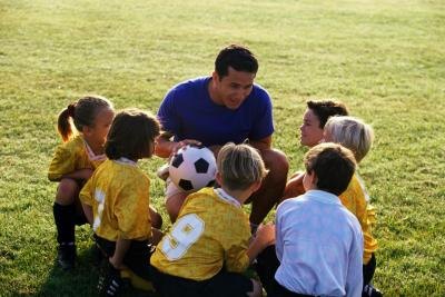 A gym teacher explains the game of soccer to young children on a field.