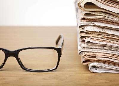 A pair of black eyeglass frames and a stack of newspapers on a desk.