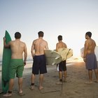 Three men sitting on wet sand by surfboards, rear view