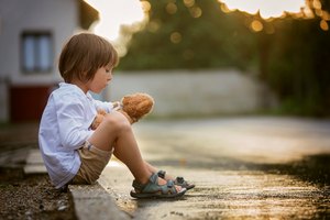 Sad little boy, sitting on the street in the rain, hugging his teddy bear, summertime on sunset