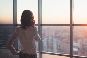 Rear view portrait of young worker speaking using cell phone, looking out the window. Female having business call, busy at her workplace in evening.