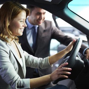 A woman sitting in the driver's seat of a new car with a salesman standing next to her